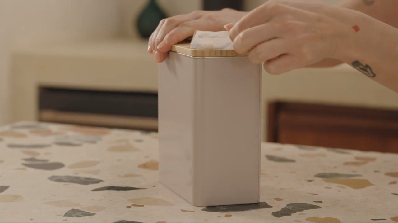 A patient lifting the day’s supplement sachet from its tin at a kitchen table.